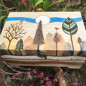 May include: A wooden box with a hand-painted landscape design. The lid features a blue sky with a white moon, mountains, trees, and birds. The box has a metal clasp and is made of light-coloured wood.