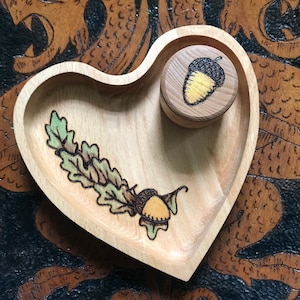 May include: A heart-shaped wooden dish with a burnt design of an acorn and leaves. The dish has a smaller round wooden dish with a burnt acorn design inside.