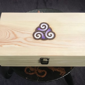 May include: A rectangular wooden box with a light finish. The box features a decorative emblem of three swirling shapes in purple, white, and brown. A metal clasp secures the lid. The box is sitting on a dark surface.