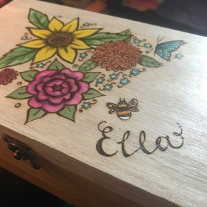 May include: A wooden box with a whitewash finish, decorated with a woodburned floral design. The design includes a sunflower, a rose, a chrysanthemum, a butterfly, and a bee. The name "Ella" is also woodburned on the box.