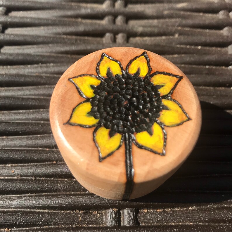 May include: A small, round, light-coloured wooden object featuring a hand-burned and painted sunflower design on its top surface. The sunflower has bright yellow petals and a dark, textured centre, with a single stem extending downwards.