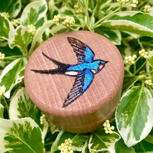 May include: A circular wooden box featuring a blue and black swallow design. The bird is depicted mid-flight on the lid, which has a visible wood grain. The box is set against a backdrop of green and white variegated leaves.