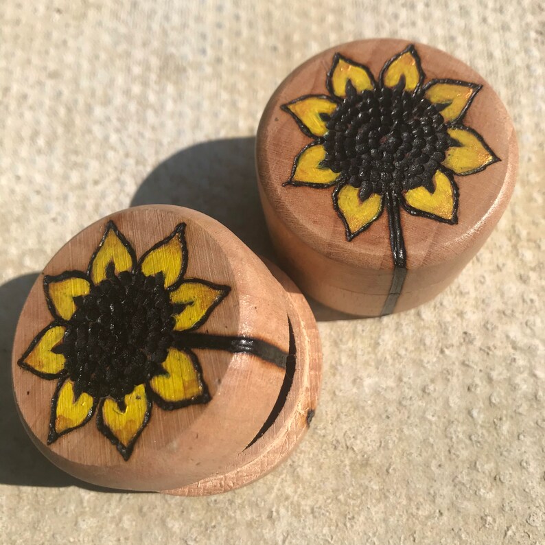 May include: Two round wooden containers with lids, each featuring a hand-burned and painted sunflower design on top. The sunflowers have dark brown centres and yellow petals, with a dark line extending from the base of each flower.
