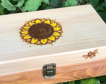 Puede incluir: Una caja de madera de color claro con un diseño de girasol en la tapa. El girasol es amarillo y marrón, con un centro detallado. Una pequeña abeja también está grabada en la madera. La caja tiene un cierre metálico.