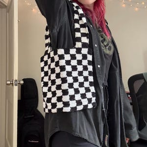 May include: A handmade crochet tote bag with a black and white checkerboard pattern. The bag is held up by a person. The bag is made of a soft yarn and has a simple, functional design. The bag is perfect for carrying books, groceries, or other items.