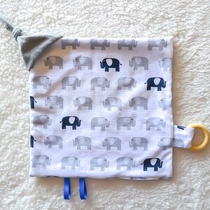 May include: A white baby blanket with a gray elephant pattern and a blue ribbon. The blanket has a gray fabric tag with a knot and a wooden teething ring attached.