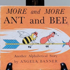 Op de afbeelding: Een oranje boekomslag met de titel "MORE and MORE ANT and BEE" in zwarte tekst. De omslag toont een illustratie van een mier en een bij die op een pijl rijden. De tekst "Another Alphabetical Story by ANGELA BANNER" is onder de illustratie gedrukt.