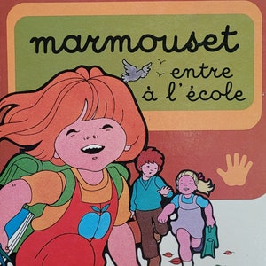 May include: A vintage children's book cover featuring a girl with red hair and a red and orange outfit walking with two other children. The book title is "marmouset entre à l'école" and the author is "dina-k. tourneur". The cover is illustrated in a cartoon style with a bright color palette.