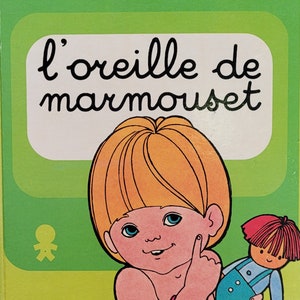 May include: A green book with the title "l'oreille de marmouset" by dina-k. tourneur. The cover features a cartoon illustration of a child with blonde hair and a doll.