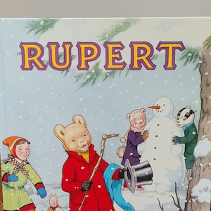 Könnte beinhalten: Ein Vintage-Kinderbuchcover mit Rupert Bär in einem roten Mantel und gelben Hosen, der in einer Schneelandschaft mit einem Schneemann, einem Schwein, einem Dachs und einem grünen Drachen steht. Der Buchtitel lautet "Rupert" in gelben Buchstaben mit violetter Umrandung.