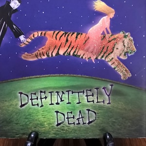 May include: A book cover for "Definitely Dead" by Charlaine Harris. The cover features a purple background with the author's name in gold. A woman rides a glittery tiger, and a vampire is present. The title is in green.