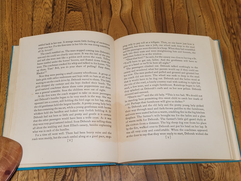 K&ouml;nnte beinhalten: Ein altes Buch, das auf den Seiten 8 und 9 aufgeschlagen ist und eine Schwarzwei&szlig;illustration einer von Pferden gezogenen Kutsche mit Passagieren zeigt. Der Text auf der Seite beschreibt eine Reise in einer Pferdekutsche.
