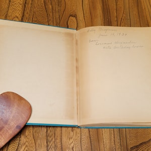 K&ouml;nnte beinhalten: Ein ge&ouml;ffnetes Buch mit einem blauen Einband. Die Innenseiten sind leer, bis auf eine handschriftliche Widmung, die lautet: "Dolly Chapman 14. Juni 1939 von Doris und Alexander Mit Geburtstagsliebe."