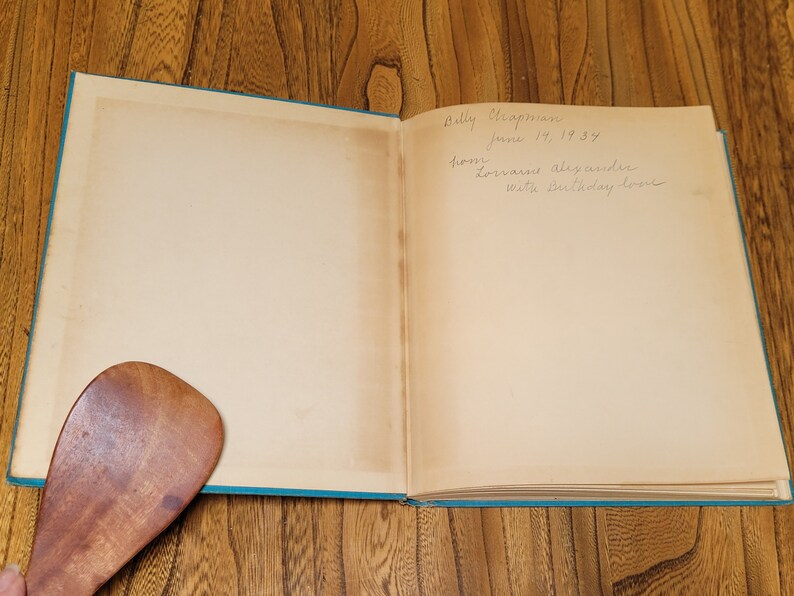 K&ouml;nnte beinhalten: Ein ge&ouml;ffnetes Buch mit einem blauen Einband. Die Innenseiten sind leer, bis auf eine handschriftliche Widmung, die lautet: "Dolly Chapman 14. Juni 1939 von Doris und Alexander Mit Geburtstagsliebe."