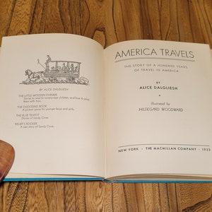 K&ouml;nnte beinhalten: Ein ge&ouml;ffnetes Buch mit einem blauen Einband. Das Buch tr&auml;gt den Titel "America Travels: The Story of a Hundred Years of Travel in America" von Alice Dalgliesh, illustriert von Hildegarde Woodward. Das Buch wurde 1933 von The Macmillan Company ver&ouml;ffentlicht.