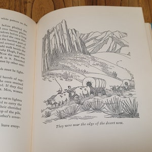K&ouml;nnte beinhalten: Eine Schwarzwei&szlig;illustration einer Gruppe von Planwagen, die durch eine W&uuml;stenlandschaft fahren. Die Wagen werden von Ochsen gezogen und sind von hohen, d&uuml;nnen W&uuml;stenpflanzen umgeben. Der Text "They were near the edge of the desert now." ist unten im Bild gedruckt.