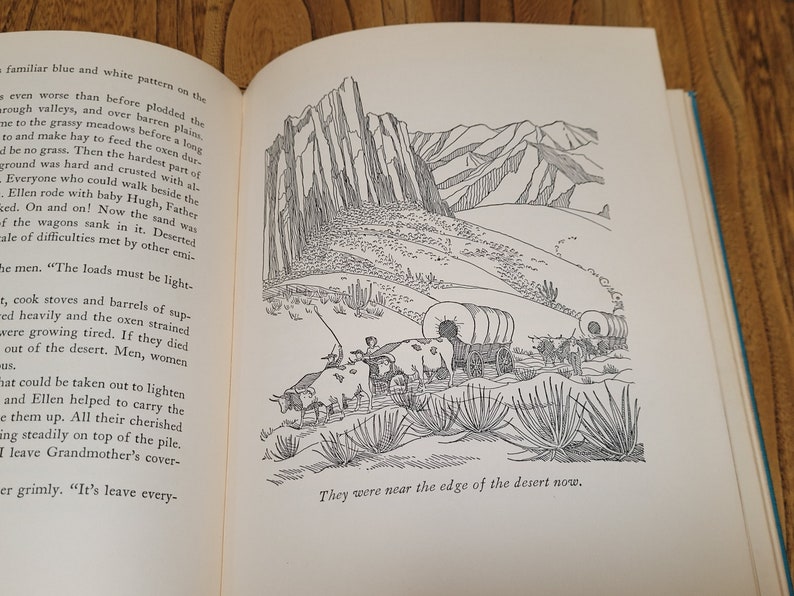 K&ouml;nnte beinhalten: Eine Schwarzwei&szlig;illustration einer Gruppe von Planwagen, die durch eine W&uuml;stenlandschaft fahren. Die Wagen werden von Ochsen gezogen und sind von hohen, d&uuml;nnen W&uuml;stenpflanzen umgeben. Der Text "They were near the edge of the desert now." ist unten im Bild gedruckt.