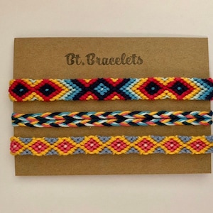 Pode incluir: Três pulseiras de amizade coloridas com padrões geométricos em tons de vermelho, azul, amarelo e rosa. As pulseiras são exibidas em um cartão marrom com o texto "Bt. Bracelets".