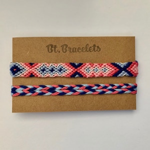 Op de afbeelding: Twee handgemaakte vriendschapsarmbanden op een bruine kaart. De bovenste armband heeft een geometrisch patroon in roze, blauw en wit. De onderste armband is gevlochten met roze, lichtblauwe en marineblauwe draden. De kaart is gelabeld met "Bt. Bracelets."