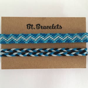 Puede incluir: Dos pulseras de la amistad en una tarjeta marrón con el texto "Bt. Bracelets". La pulsera superior es azul, blanca y gris con un patrón de chevron. La pulsera inferior es negra, azul y blanca con un patrón trenzado.