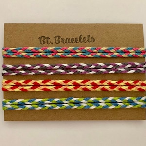 Op de afbeelding: Gevlochten vriendschapsarmbanden in diverse kleuren, gepresenteerd op een bruine kaart. De armbanden hebben een geweven ontwerp met kleuren als roze, blauw, paars, rood, geel, groen en wit. De kaart is voorzien van de tekst "Bt. Bracelets".
