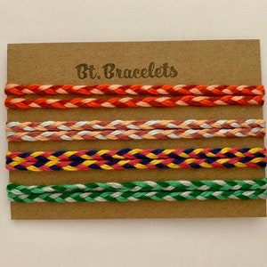 Op de afbeelding: Set van vier gevlochten armbanden in verschillende kleuren, waaronder rood, oranje, roze, wit, geel, blauw, groen en donkerblauw. De armbanden worden getoond op een bruine kaart met de tekst "Bt. Bracelets".