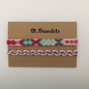 Może przedstawiać: Dwa bransoletki przyjaźni na brązowej kartce z tekstem "Bt. Bracelets". Górna bransoletka to wzór tkany w kolorze różowym, zielonym i białym. Dolna bransoletka to wzór pleciony w kolorze różowym, białym i turkusowym.