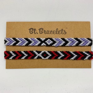 Op de afbeelding: Twee vriendschapsarmbanden op een bruine kaart met de tekst "Bt. Bracelets". De bovenste armband is paars, wit en zwart. De onderste armband is rood, wit en zwart.