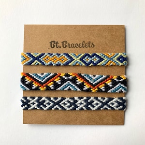 Könnte beinhalten: Drei Freundschaftsarmbänder mit geometrischen Mustern in Blau, Gelb, Orange und Weiß. Die Armbänder werden auf einer braunen Karte mit dem Text "Bt. Bracelets" präsentiert.