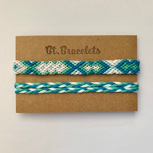 Puede incluir: Dos pulseras de amistad en una tarjeta marrón con el texto "Bt. Bracelets". La pulsera superior es un patrón de chevron blanco, azul y verde. La pulsera inferior es un patrón trenzado blanco, azul y verde.