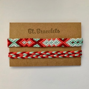 Könnte beinhalten: Zwei Freundschaftsarmbänder auf einer braunen Karte mit dem Text "Bt. Bracelets". Das obere Armband ist ein rotes, weißes und hellblaues gewebtes Muster. Das untere Armband ist ein rotes und hellblaues geflochtenes Muster.