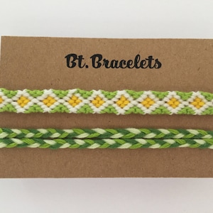 May include: Two friendship bracelets on a brown card with the text "Bt. Bracelets". The top bracelet is green and white with yellow squares. The bottom bracelet is green and white.
