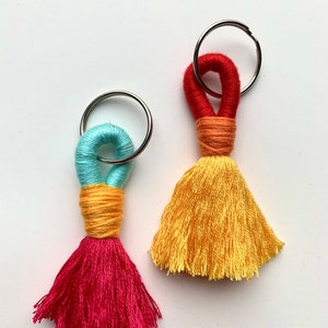 Puede incluir: Dos llaveros de borla coloridos con anillos de metal. Un llavero es rojo, amarillo y turquesa. El otro llavero es amarillo, naranja y rojo.