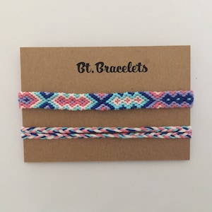 May include: Two friendship bracelets on a brown card with the text "Bt. Bracelets". The top bracelet is a woven pattern with pink, blue, and white thread. The bottom bracelet is a braided pattern with pink, blue, and white thread.