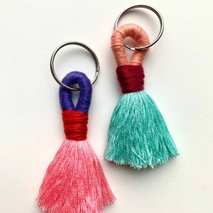 Puede incluir: Dos llaveros de borla coloridos con anillos de llave plateados. El llavero de la izquierda tiene una borla rosa con detalles en rojo, blanco y azul. El llavero de la derecha tiene una borla azul verdosa con detalles en rosa, rojo y morado.