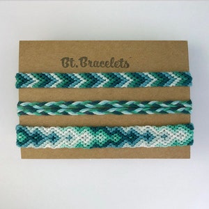 Op de afbeelding: Drie vriendschapsarmbanden in tinten blauw en groen. De armbanden zijn gemaakt met een chevron-patroon en een gevlochten patroon. De armbanden worden getoond op een bruine kaart met de tekst "Bt. Bracelets".