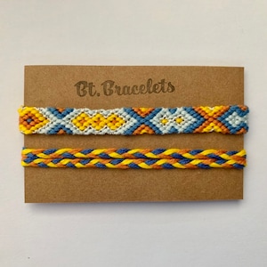Op de afbeelding: Twee vriendschapsarmbanden op een bruine kaart met de tekst "Bt. Bracelets". De bovenste armband is een geweven patroon in blauw, wit, geel en oranje. De onderste armband is een gevlochten patroon in blauw, geel en oranje.