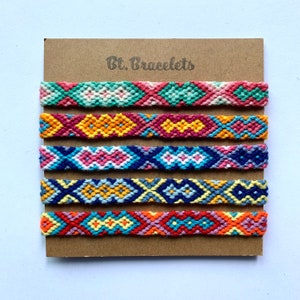 Op de afbeelding: Set van zes kleurrijke vriendschapsarmbanden met geometrische patronen. De armbanden zijn gemaakt van kleurrijk borduurgaren en zijn gerangschikt in een rij op een bruine kaart met de tekst "Bt. Bracelets".