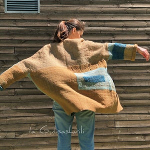 Puede incluir: Una mujer lleva un cárdigan largo de patchwork marrón, azul y verde con unos vaqueros azules de denim. El cárdigan tiene un corte holgado y está hecho de una tela suave y texturizada. El texto "la Gulisashvili" es visible en la parte inferior de la imagen.