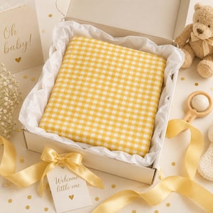 May include: A square yellow and white gingham patterned item nestled in a white gift box lined with tissue paper. The box includes a tan teddy bear, a wooden rattle, a card with the text Oh baby, and a tag with the text Welcome little one.