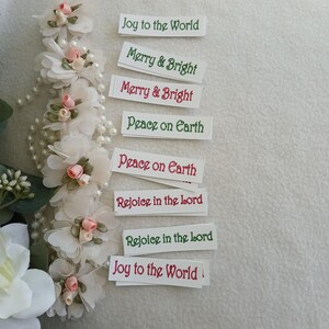 May include: A vertical garland of white fabric flowers with small pink buds and pearl beads, accompanied by eight rectangular paper tags featuring the printed phrases Joy to the World, Merry & Bright, Peace on Earth, and Rejoice in the Lord.