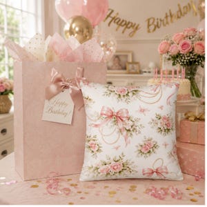 Puede incluir: Un cojín cuadrado con fondo blanco y un patrón repetitivo de rosas rosas, lazos de color rosa claro y cadenas doradas. El cojín está colocado junto a una bolsa de regalo de color rosa claro con un lazo a juego y una etiqueta de Happy Birthday.
