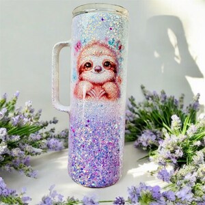 May include: A white and purple glitter tumbler with a sloth design. The sloth is brown and has a pink nose and a white flower crown. The tumbler has a handle and is made of stainless steel.