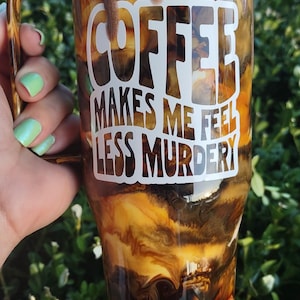 May include: A brown and gold tumbler with a white design that says "Coffee makes me feel less murdery". The tumbler has a brown drip effect on the outside.