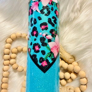 May include: A turquoise blue tumbler with a black and white leopard print design and pink flowers. The tumbler has a glitter finish.
