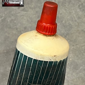 Puede incluir: Primer plano de una botella vintage con tap&oacute;n rojo. La botella tiene una parte superior color crema y un cuerpo verde oscuro con rayas plateadas verticales. El logotipo "Moonshine Antiques" est&aacute; en la esquina superior izquierda.