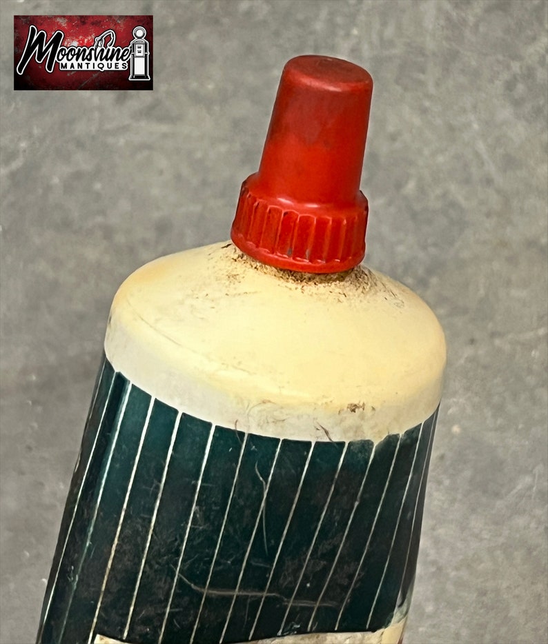 Puede incluir: Primer plano de una botella vintage con tap&oacute;n rojo. La botella tiene una parte superior color crema y un cuerpo verde oscuro con rayas plateadas verticales. El logotipo "Moonshine Antiques" est&aacute; en la esquina superior izquierda.
