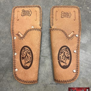 May include: Two brown leather gun holsters with decorative stitching and a cowboy and horse design. Each holster has the words "GENUINE LEATHER" stamped on the top. The holsters are from Moonshine Antiques.