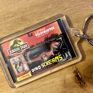 Może przedstawiać: Przezroczysty plastikowy brelok z zabawką Velociraptora z Jurassic Park w środku. Zabawka jest brązowa i zielona i ma pazur, który można poruszać. Brelok ma napis "Electronic Velociraptor - Slashing Claw!" i "Dino Screams in Electronic Sound!"