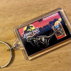 Może przedstawiać: Przezroczysty plastikowy brelok z logo Jurassic Park i czarno-zielonym obrazkiem dinozaura. Brelok zawiera tekst "Dilophosaurus" i "Dino Strike Venom Spray".